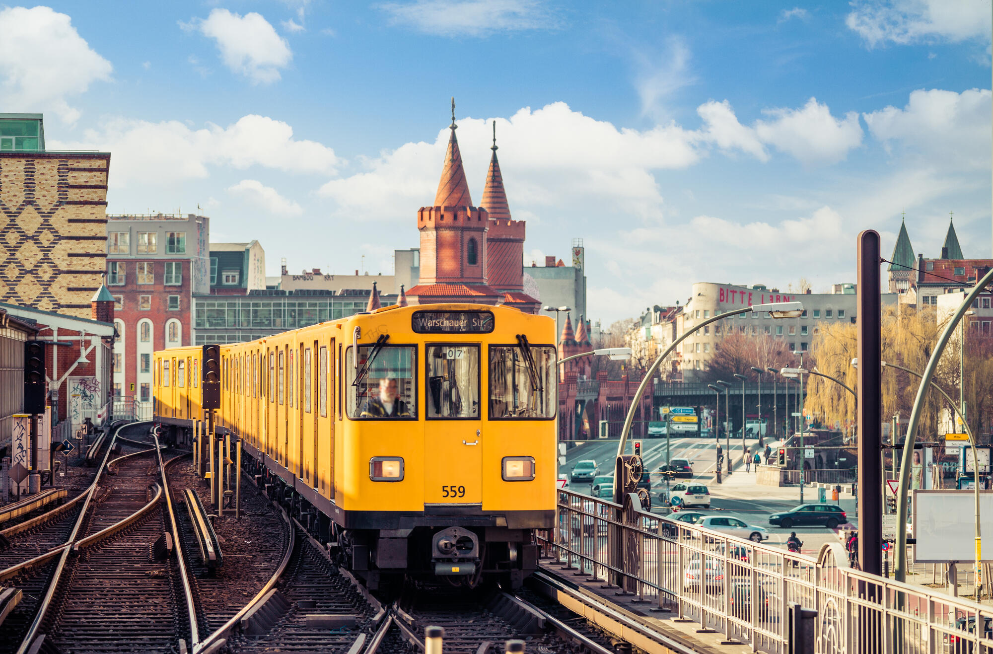 A yellow U-Bahn train passes the red towers of Oberbaum Bridge beneath bright clouds.