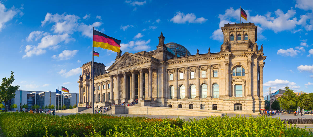 The west facade of the Reichstag with German flags flying beneath a blue sky and white clouds.