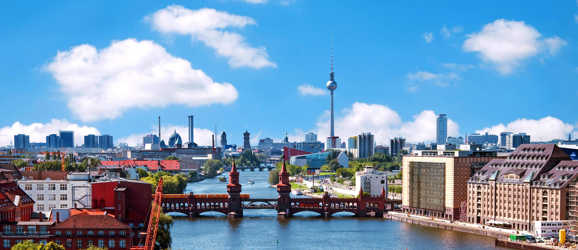 View across the Spree towards Oberbaum Bridge, with a yellow U-Bahn train and white cumulus clouds.