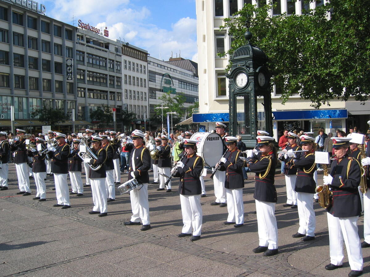 Marching band in dark uniforms and white trousers in front of the Kröpcke Clock