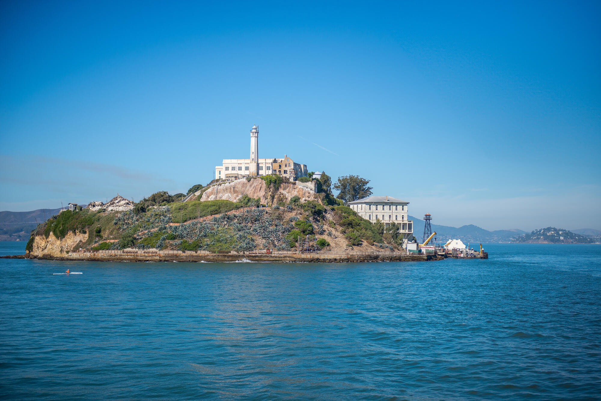 Pale buildings and the lighthouse stand on the rocky, vegetated island above the blue water of the bay.
