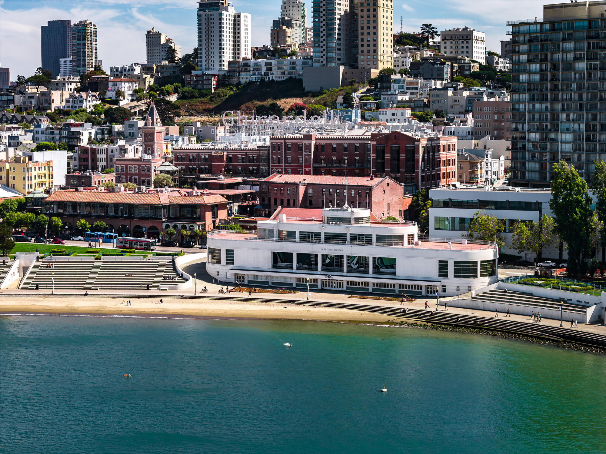 An elevated view of the curved bathhouse, seating terraces and narrow beach beneath the densely built hillside.