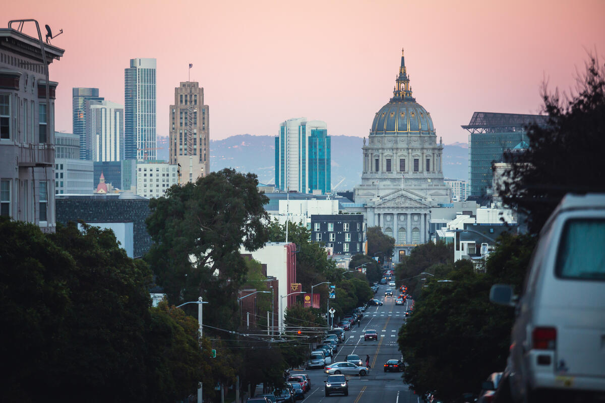A view down a hilly street between parked cars and buildings towards City Hall's dome.