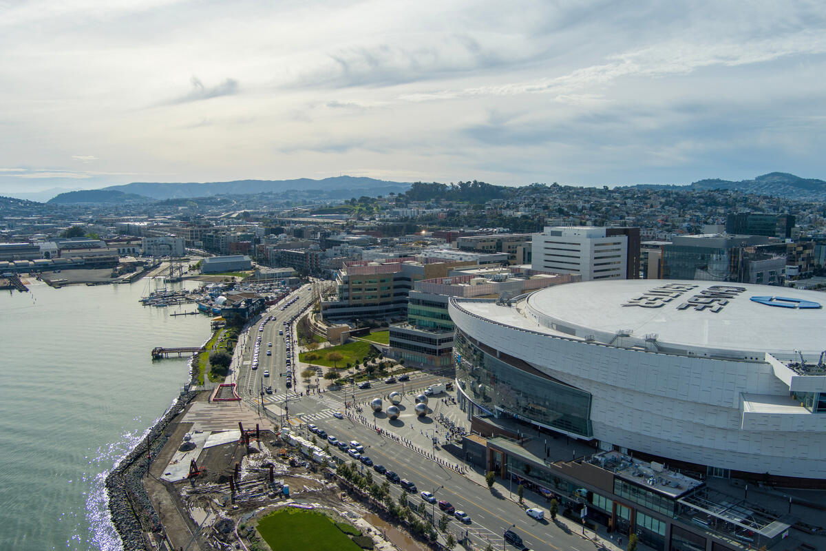 An aerial view of the curved arena, its pale outer skin and the waterfront road beside the bay.