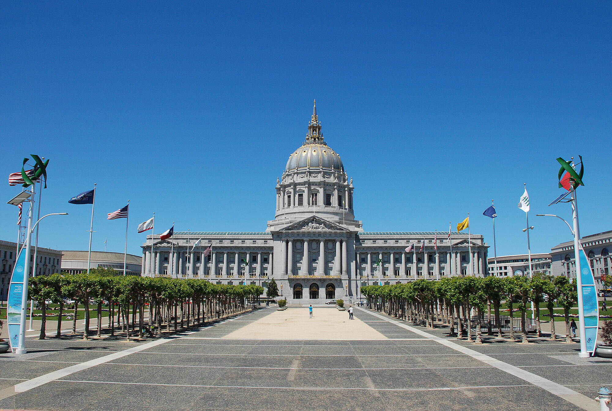 City Hall's dome and columned facade rise beyond the broad plaza, framed by rows of trees and flags.