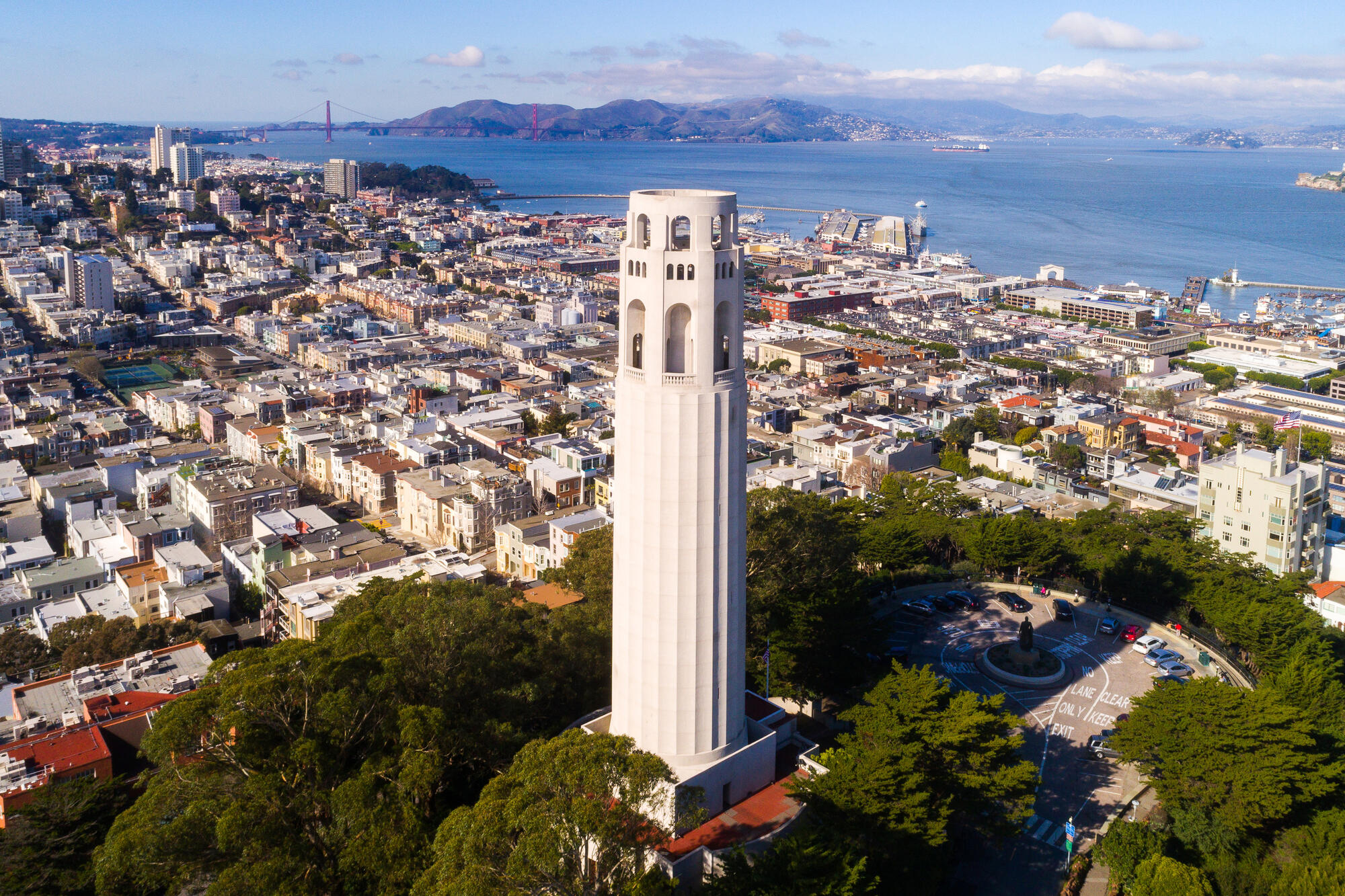 An elevated view of the pale Coit Tower among trees, dense residential streets and the bay.