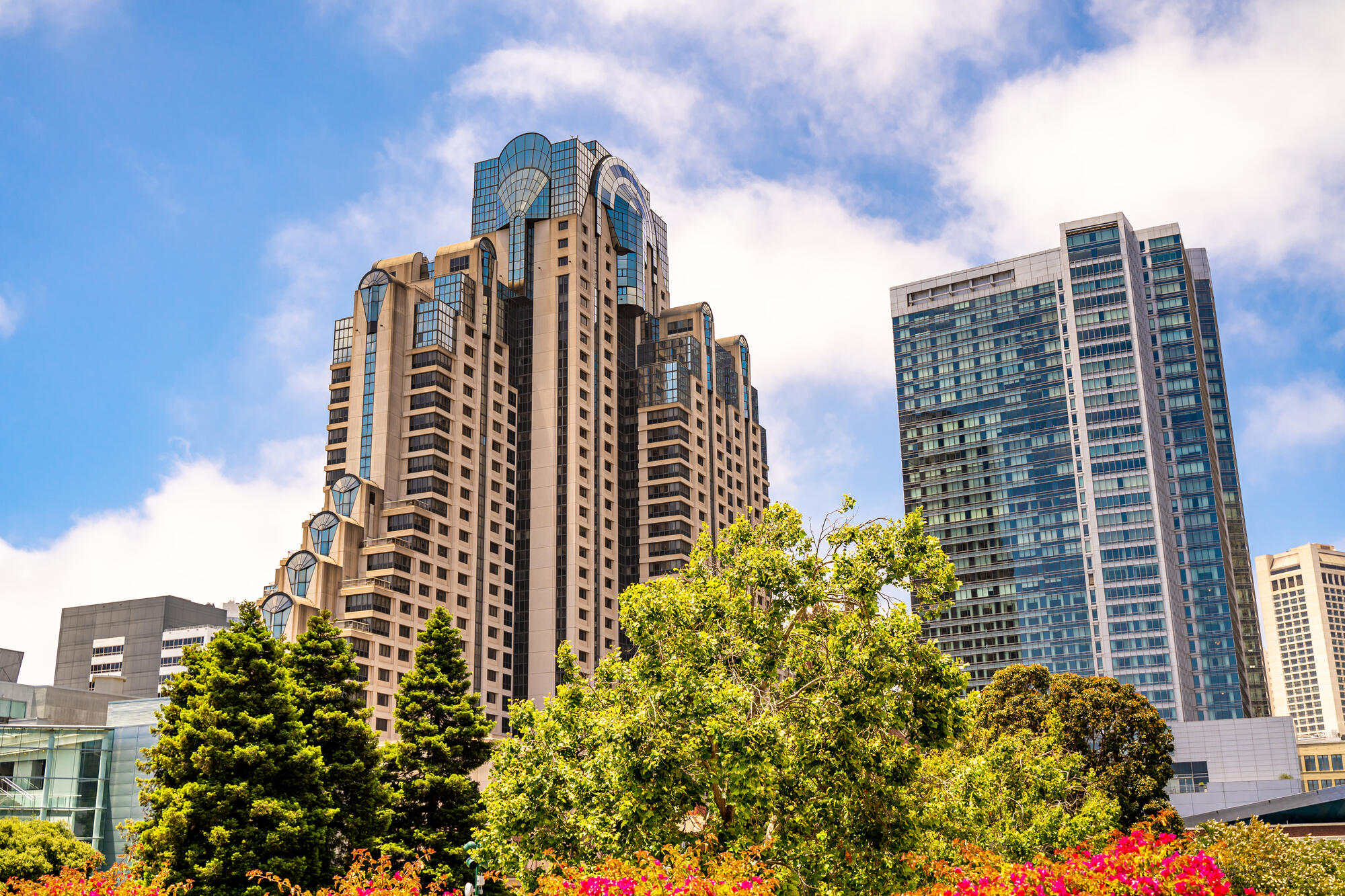 The brown hotel tower rises above trees and flower beds, with stepped projections and a fan-shaped crown.