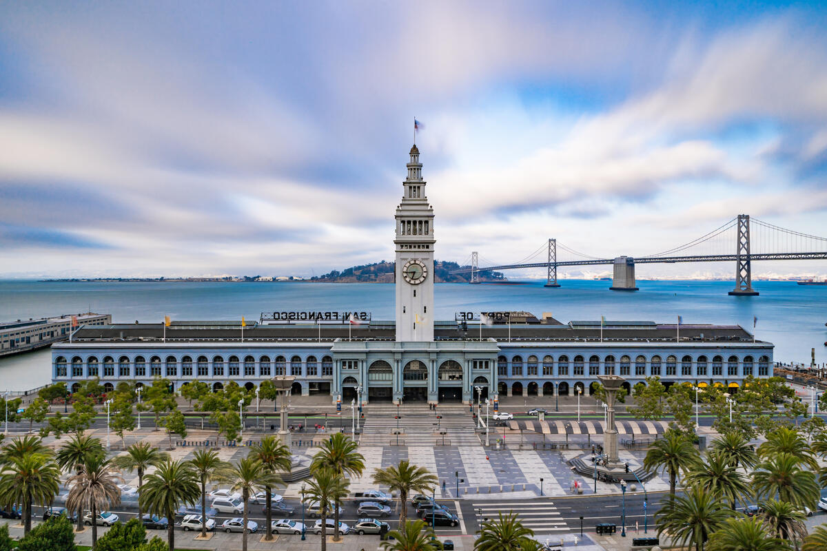 The Ferry Building's clock tower rises above its long arcaded facade, with the Bay Bridge behind.