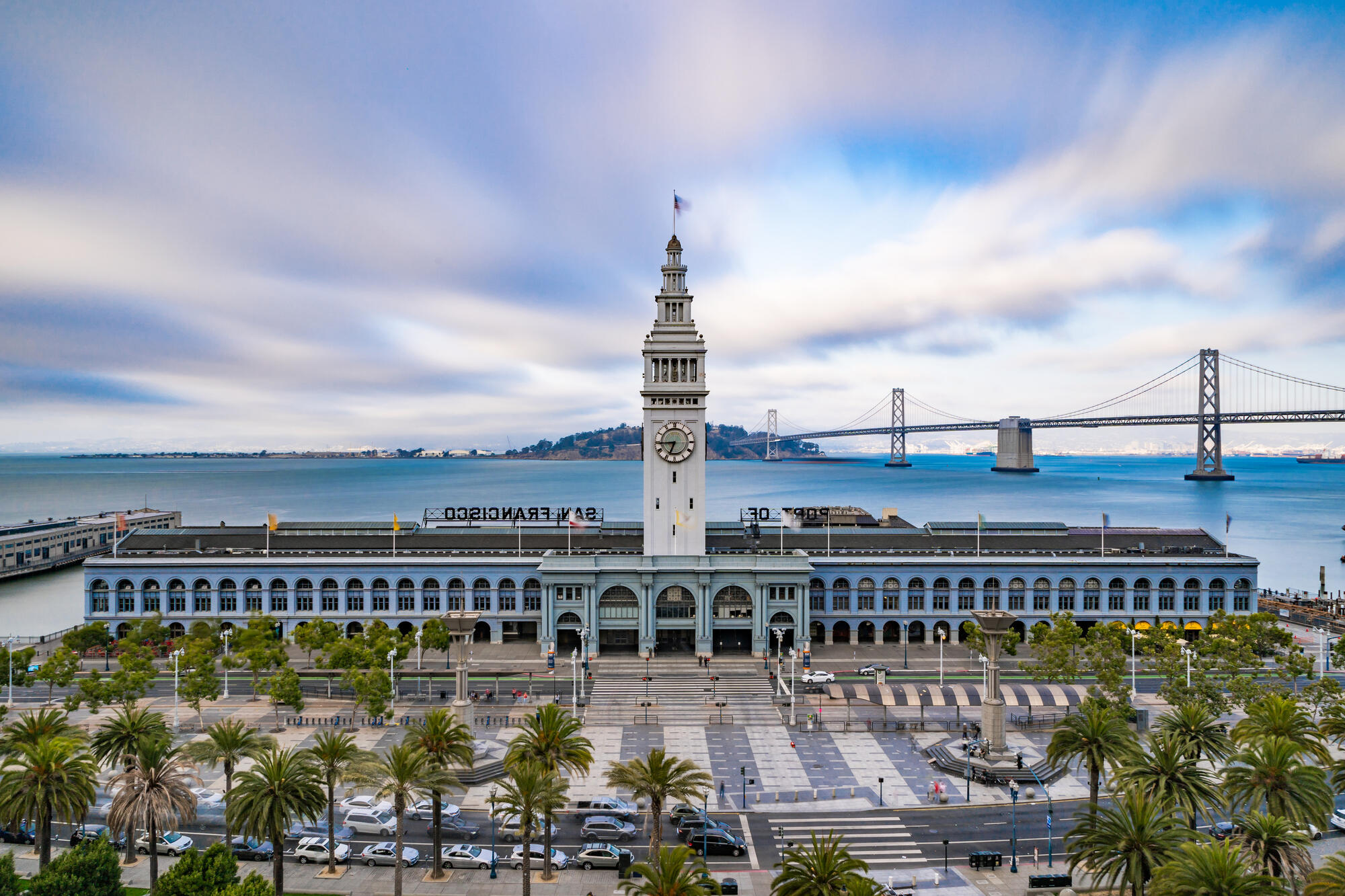 The Ferry Building's clock tower rises above its long arcaded facade, with the Bay Bridge behind.