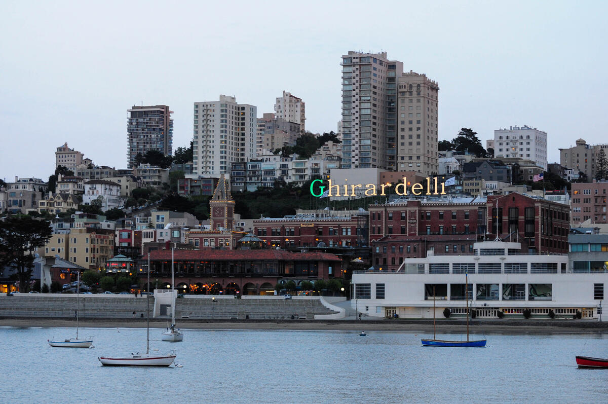 This 2012 image shows the Ghirardelli sign above brick waterfront buildings and boats in the bay.