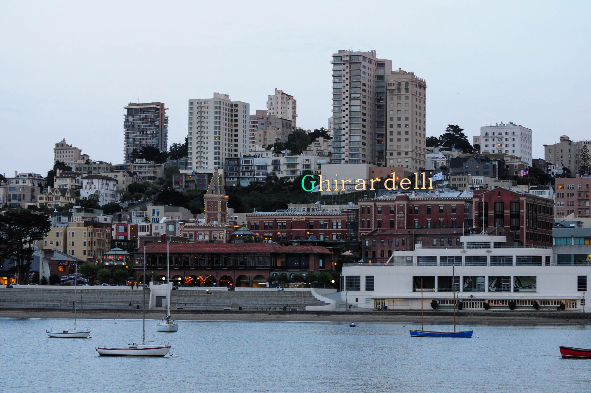 This 2012 image shows the Ghirardelli sign above brick waterfront buildings and boats in the bay.