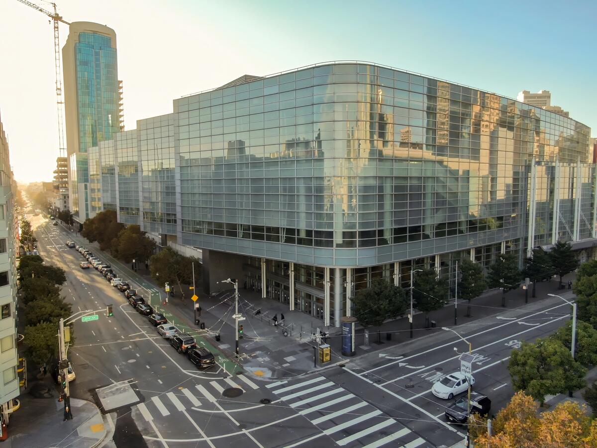 The broad convention building, with continuous glazing and pale vertical facade elements, stands at a street junction.