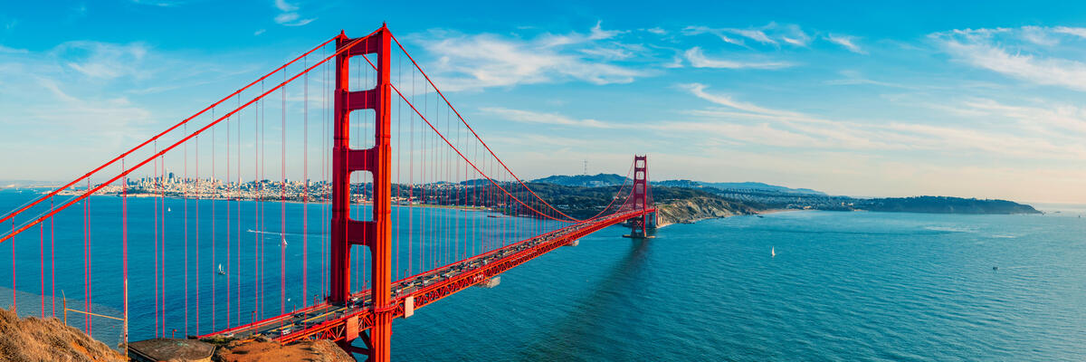 Red bridge towers and suspension cables span the blue strait, with hills on the far shore.