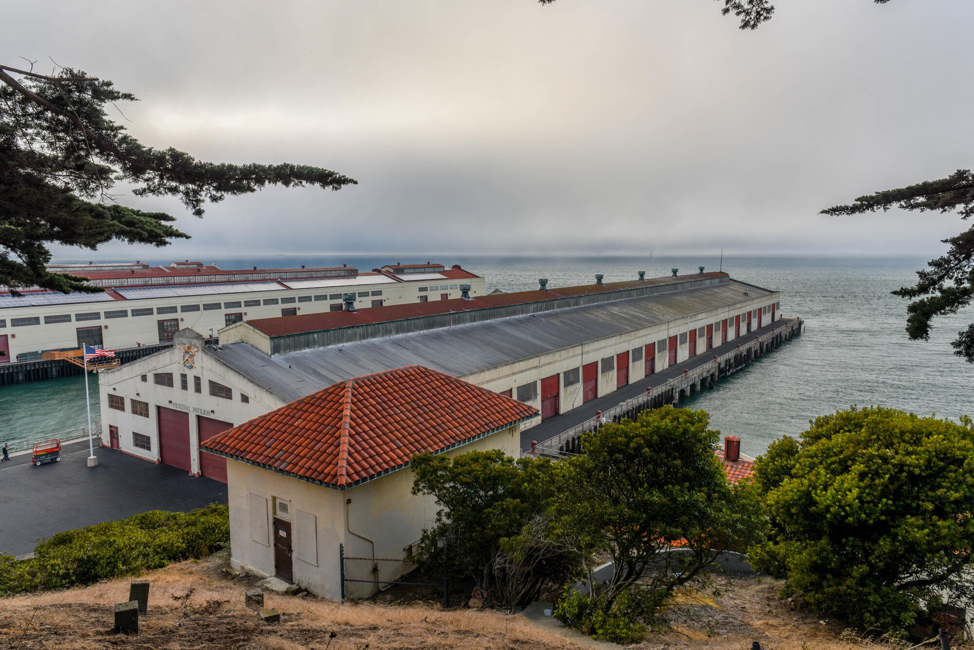 Long warehouses with pitched roofs extend into the bay beneath an overcast sky, with trees and a small tiled-roof building in the foreground.