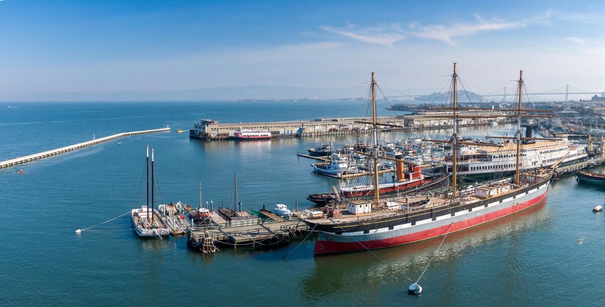 An aerial view of historic ships with tall masts and rigging at their former berths at Hyde Street Pier.