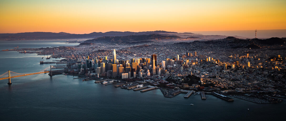 A wide aerial view of downtown between the bay and the hills in low, warm light.