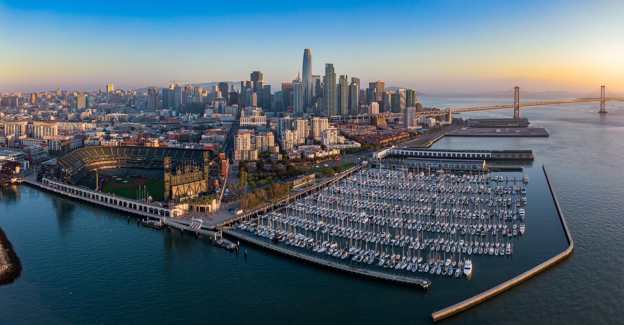 A wide aerial view of South Beach Harbor's berths, the baseball stadium and the skyline beyond.