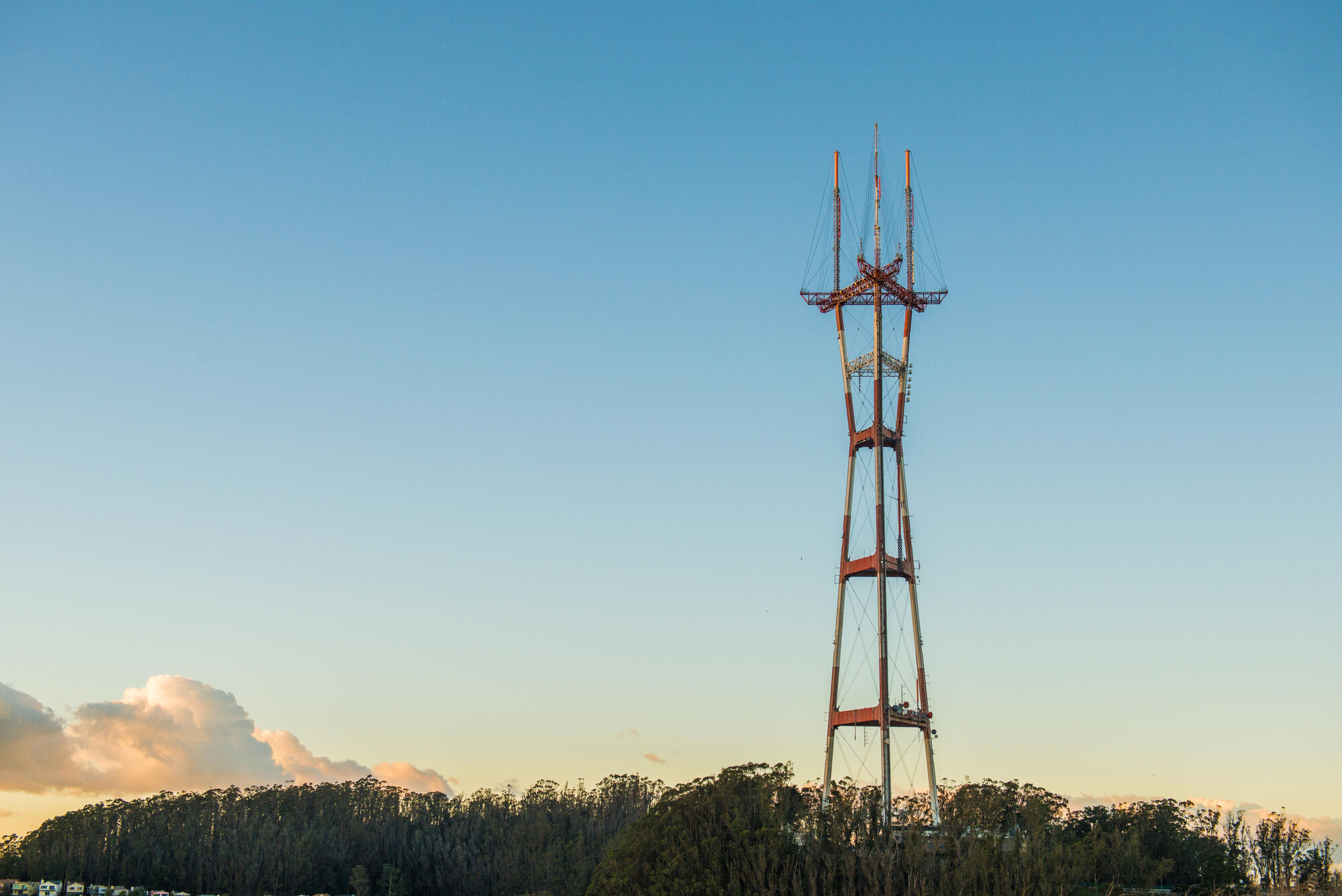 Sutro Tower's red and white steel structure rises above a dark line of trees into a pale sky.